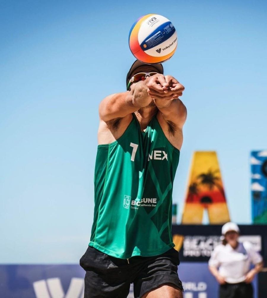 A Mexican male beach volleyball player dives for a dig on a sand court, his face focused and determined.