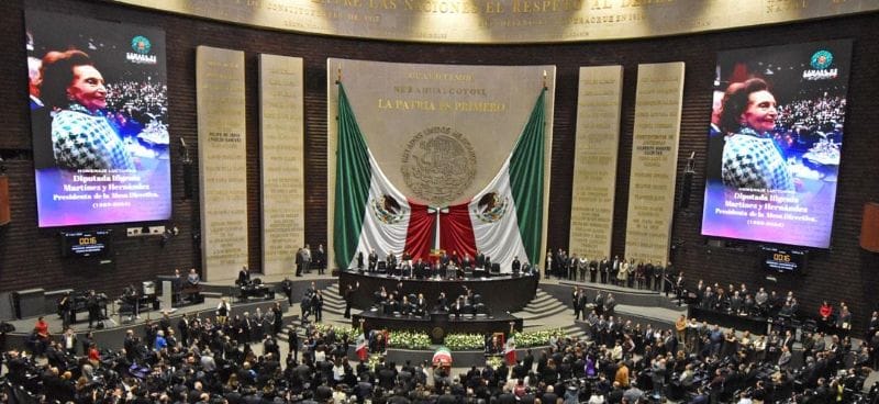 Interior view of the Mexican Chamber of Deputies filled with legislators standing in respect, with Martínez's portrait displayed prominently.