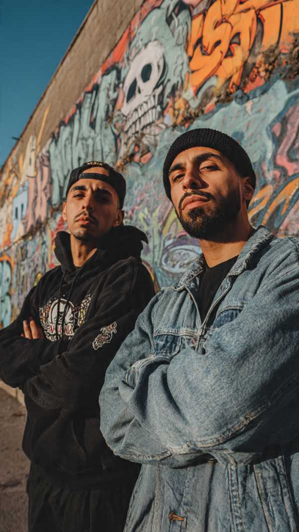 Two young Latino men pose confidently in front of a graffiti-covered wall featuring Mexican street art.