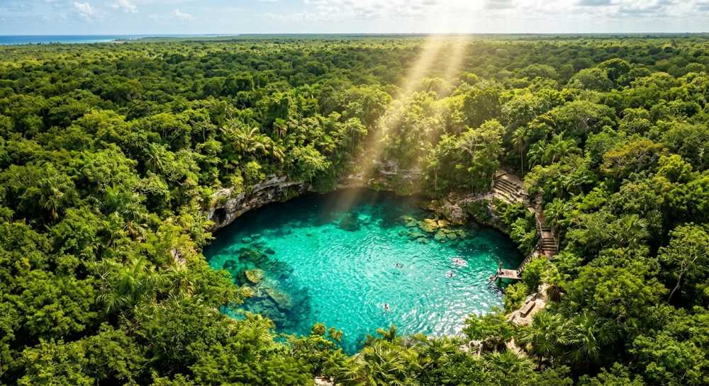 Aerial view of turquoise cenote pool surrounded by lush green jungle vegetation, sunlight streaming through trees onto crystal-clear water.