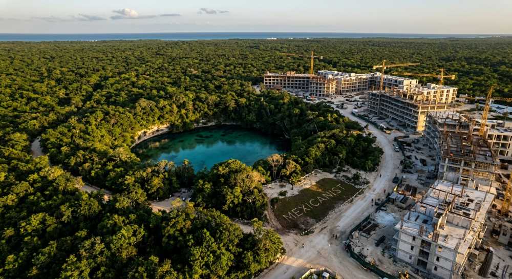 Aerial view of a cenote in the Yucatan jungle with construction cranes from hotel developments visible at the edges of the forest.