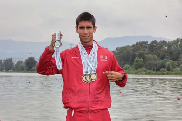 A young Latino man smiles and holds up three gold medals and one silver medal, wearing a rowing uniform.