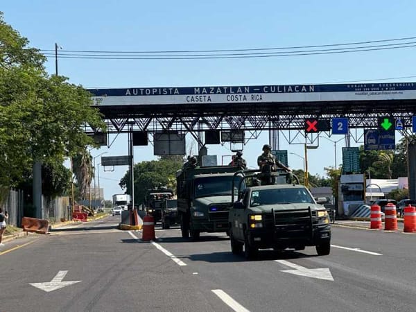 A group of Mexican Army soldiers in full combat gear standing near military vehicles at a toll booth.