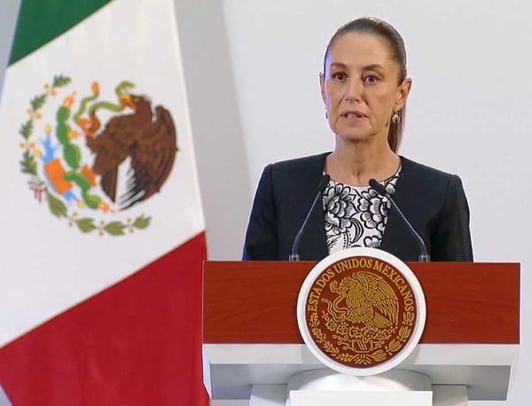 Mexican President Claudia Sheinbaum speaking at a podium with the Mexican flag visible in the background.