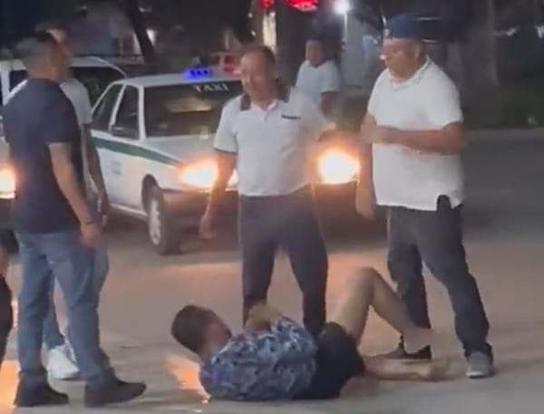 Four aggressive taxi drivers surround and kick a person lying on the ground in a Cancun street.